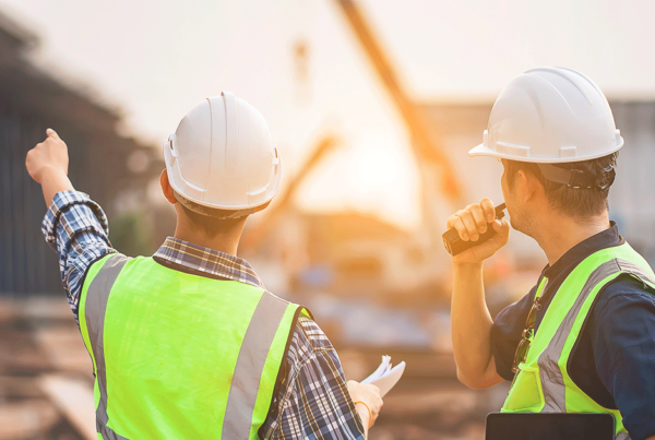 Dois trabalhadores da construção civil, utilizando capacetes e coletes refletivos, conversam durante uma vistoria de obra em um canteiro de obras.