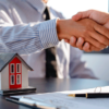 Duas pessoas apertando as mãos sobre uma mesa com uma pequena maquete de casa e documentos, indicando uma transação imobiliária