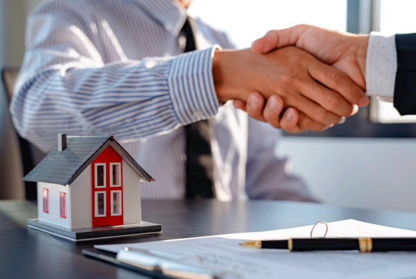 Duas pessoas apertando as mãos sobre uma mesa com uma pequena maquete de casa e documentos, indicando uma transação imobiliária